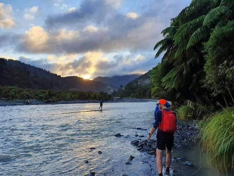 Two runners crossing a braided river at dawn, bush-clad valley walls and the sun coming up behind the ridge.